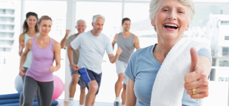 Cheerful senior woman gesturing thumbs up with people exercising in the background at fitness studio